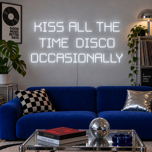 Kiss All The Time Disco Occasionally Neon Sign glowing white above a blue sofa near a disco ball.