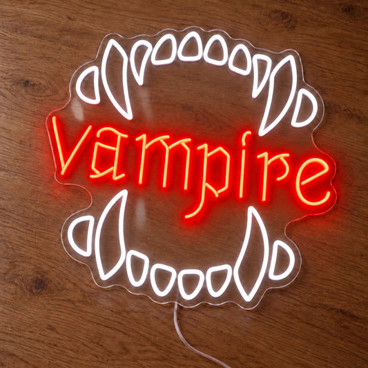Vampire Fangs Neon Sign featuring white upper and lower fangs surrounding red Vampire lettering, illuminated on clear acrylic lying on a natural oak wooden surface.