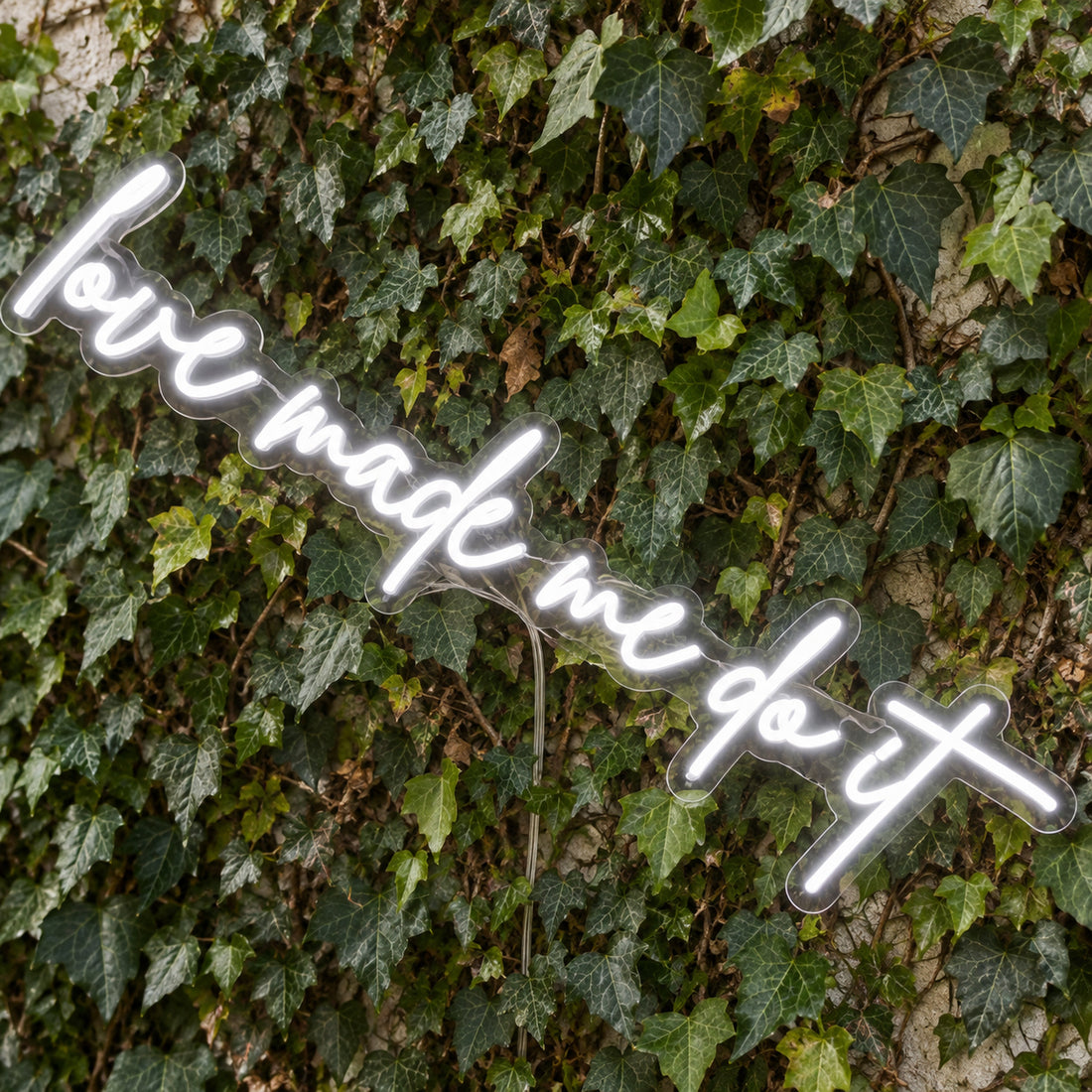 Love Made Me Do It LED Neon Light Sign in white, featuring script lettering against a green foliage wall, viewed at an angle.