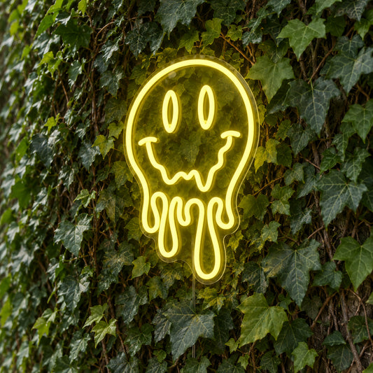 Melting Smiley Face LED Neon Light Sign in yellow, featuring dripping smiley-face outline against a green foliage wall.