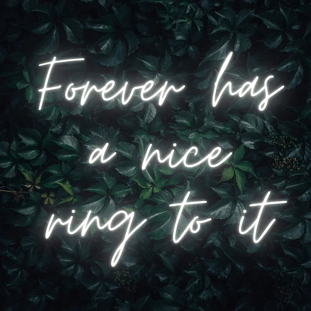 Forever Has a Nice Ring to It LED Neon Light Sign in white, featuring three lines of script lettering against a dark foliage backdrop.