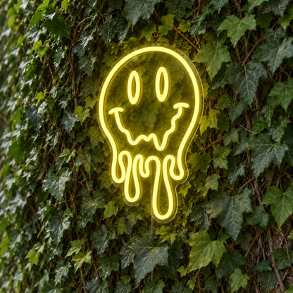 Melting Smiley Face LED Neon Light Sign in yellow, featuring dripping smiley-face outline against a green foliage wall.