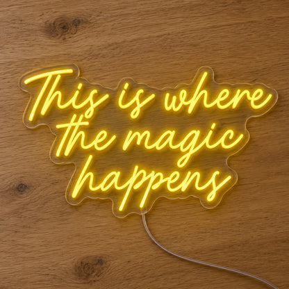 This Is Where The Magic Happens Neon Sign in yellow, featuring three lines of script lettering on a wooden surface.