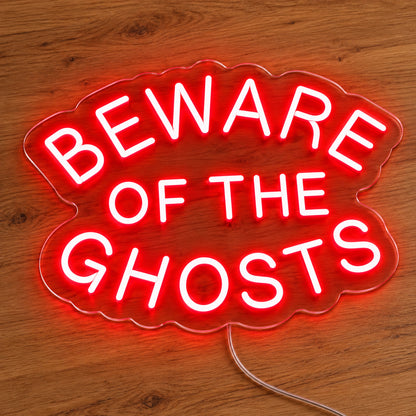 Beware of the Ghosts Neon Sign featuring Beware of the Ghosts wording arranged in curved red lettering, illuminated on clear acrylic lying on a natural oak wooden surface.