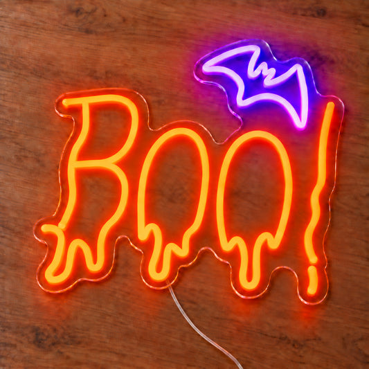 Boo Bat Neon Sign featuring orange dripping Boo lettering topped with a purple bat, illuminated on clear acrylic lying on a natural oak wooden surface.