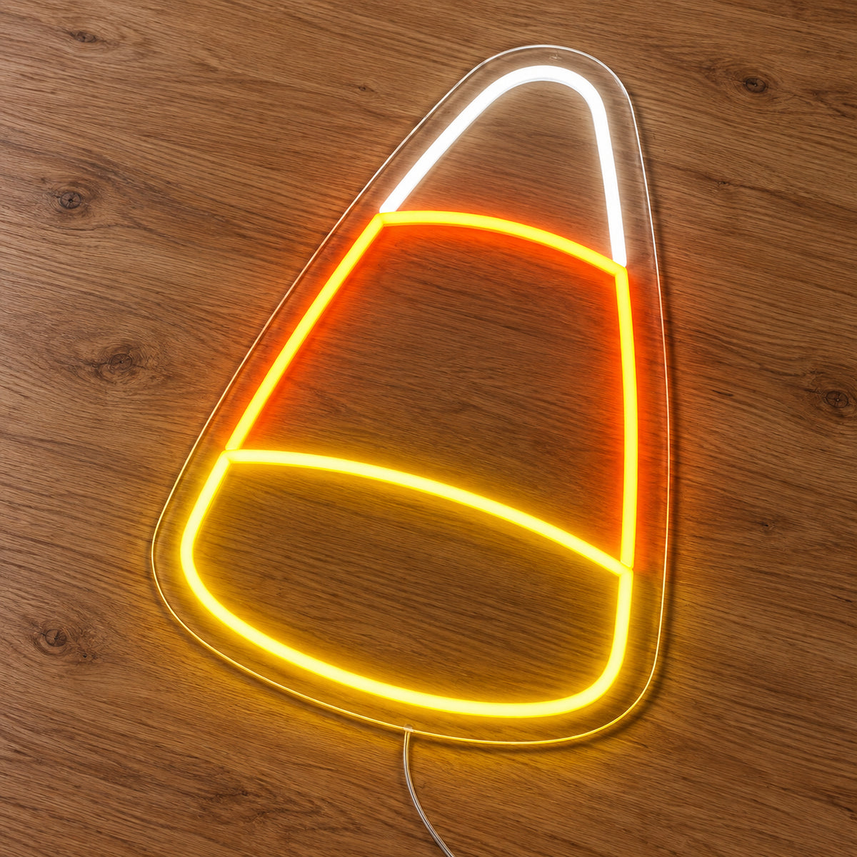 Candy Corn Neon Sign featuring a candy corn outline divided into white, orange and yellow sections, illuminated on clear acrylic lying on a natural oak wooden surface.