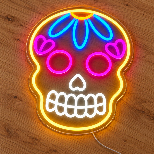 Colourful Sugar Skull Neon Sign featuring a yellow skull outline with pink eyes, a blue forehead flower and white teeth, illuminated on clear acrylic lying on a natural oak wooden surface.