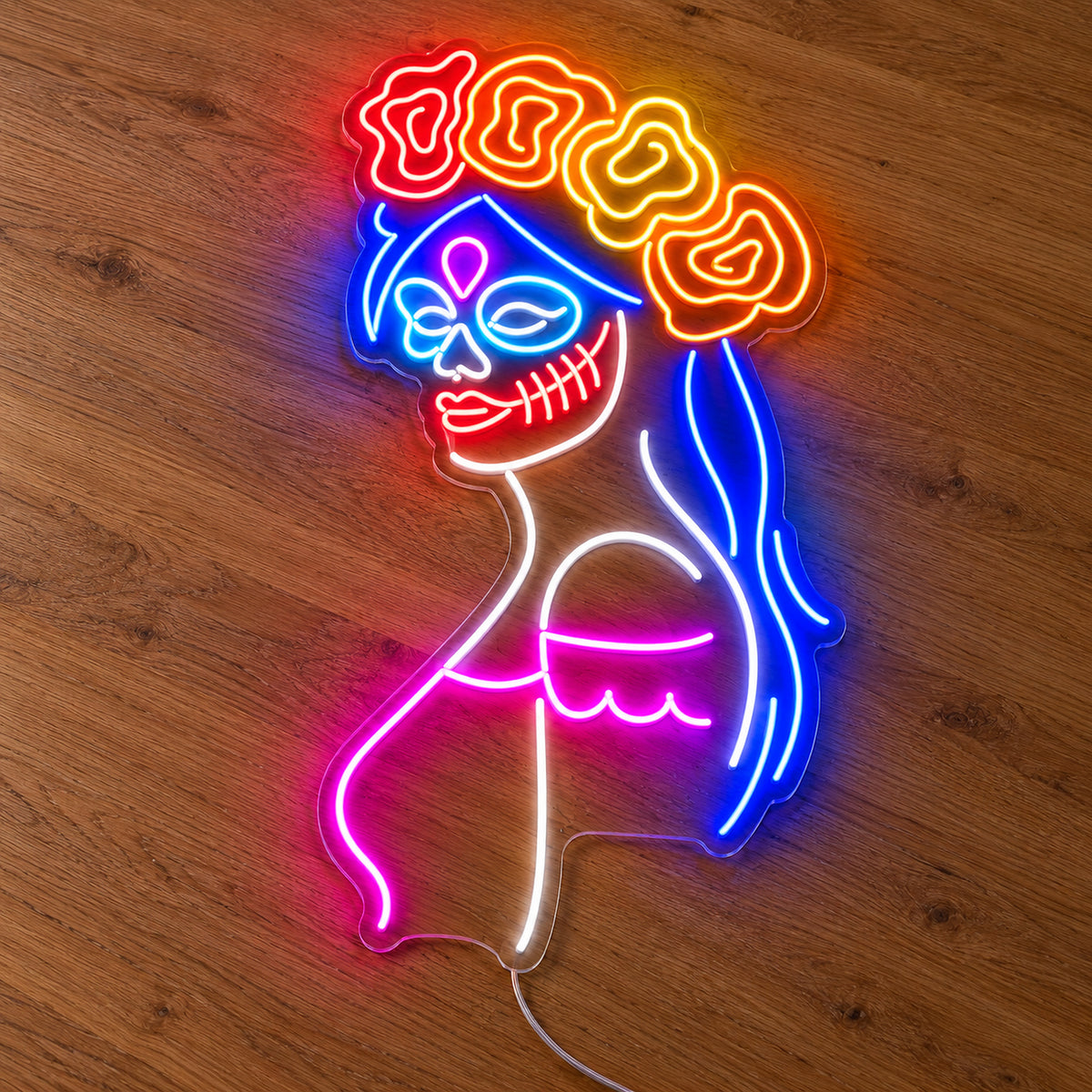 Day of the Dead Woman Neon Sign featuring a woman in profile with a decorative skull face, blue hair, a floral crown and pink accents, illuminated on clear acrylic lying on a natural oak wooden surface.