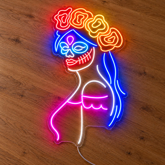 Day of the Dead Woman Neon Sign featuring a woman in profile with a decorative skull face, blue hair, a floral crown and pink accents, illuminated on clear acrylic lying on a natural oak wooden surface.