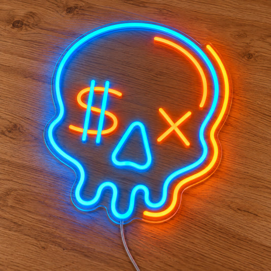 Dollar Sign Skull Neon Sign featuring a blue and orange skull with a dollar sign eye and an X shaped eye, illuminated on clear acrylic lying on a natural oak wooden surface.