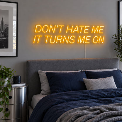 Don't Hate Me It Turns Me On Neon Sign glowing gold above a bed with dark blue bedding.