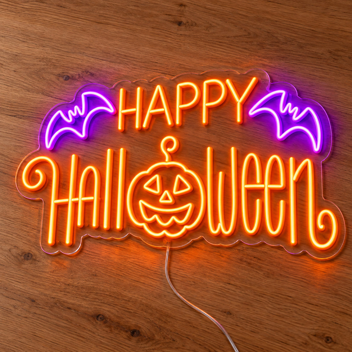 Happy Halloween Pumpkin and Bats Neon Sign featuring orange Happy Halloween lettering, a smiling pumpkin face and purple bats, illuminated on clear acrylic lying on a natural oak wooden surface.