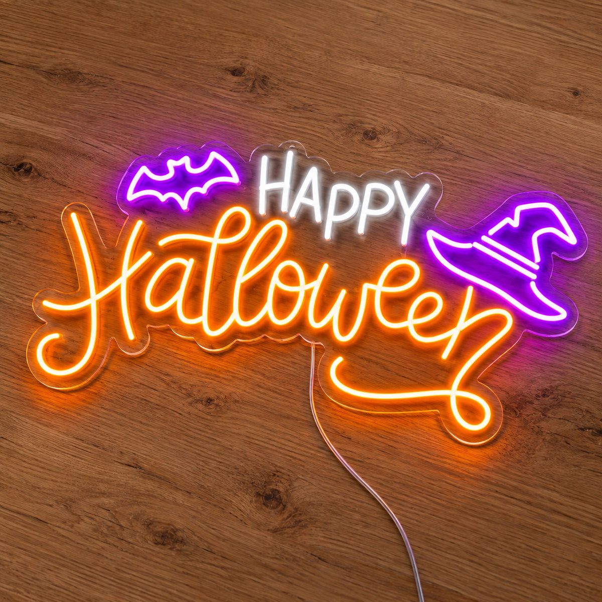 Happy Halloween Witch Hat Neon Sign featuring white Happy lettering, orange Halloween script, a purple bat and a purple witch hat, illuminated on clear acrylic lying on a natural oak wooden surface.