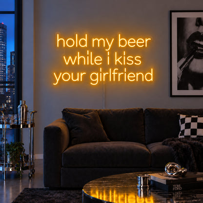Hold My Beer While I Kiss Your Girlfriend Neon Sign glowing gold above a burgundy sofa.