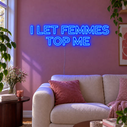 I Let Femmes Top Me Neon Sign glowing blue in outlined lettering above a cream chair.