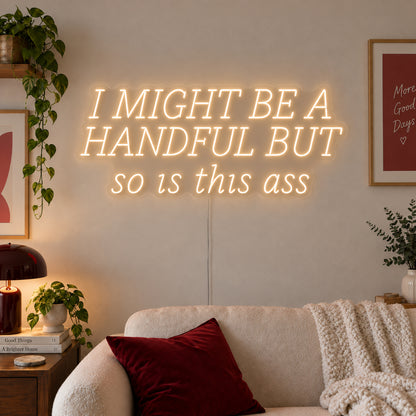 Warm White version of the I Might Be A Handful But So Is This Ass Neon Sign, shown in wide lettering above a cream sofa.