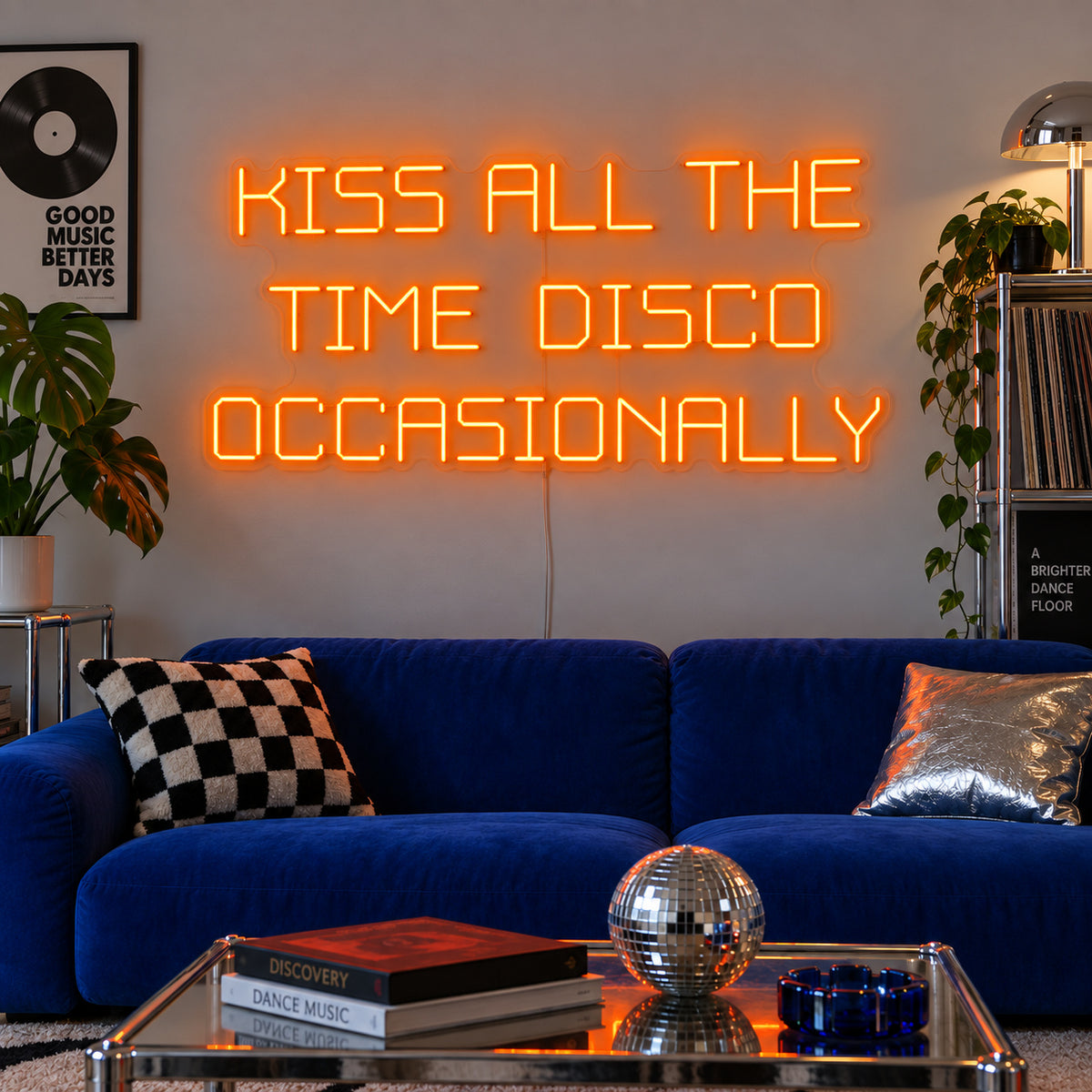 Orange version of the Kiss All The Time Disco Occasionally Neon Sign, shown above a blue sofa near a disco ball.