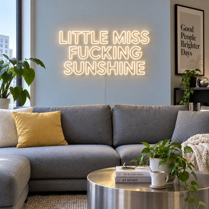 Warm White version of the Little Miss Fucking Sunshine Neon Sign, shown above a grey sofa with yellow cushions.