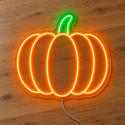 Orange Pumpkin Neon Sign featuring an orange pumpkin outline with curved segments and a green stem, illuminated on clear acrylic lying on a natural oak wooden surface.