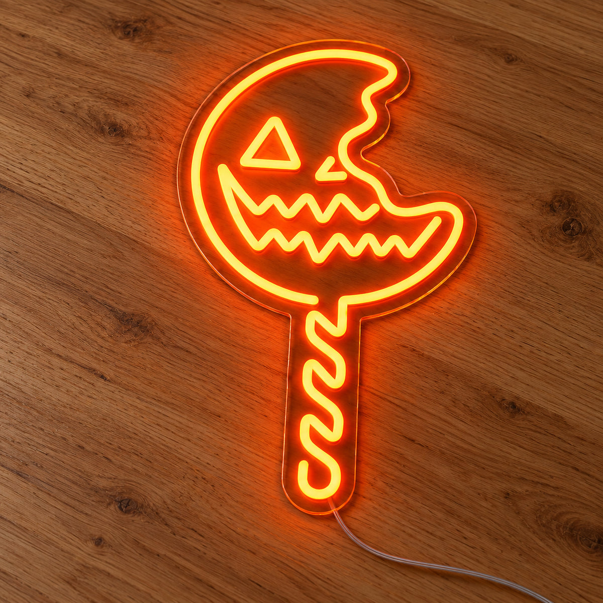 Pumpkin Lollipop Neon Sign featuring an orange jack o lantern lollipop with a bite shaped edge and a striped stick, illuminated on clear acrylic lying on a natural oak wooden surface.