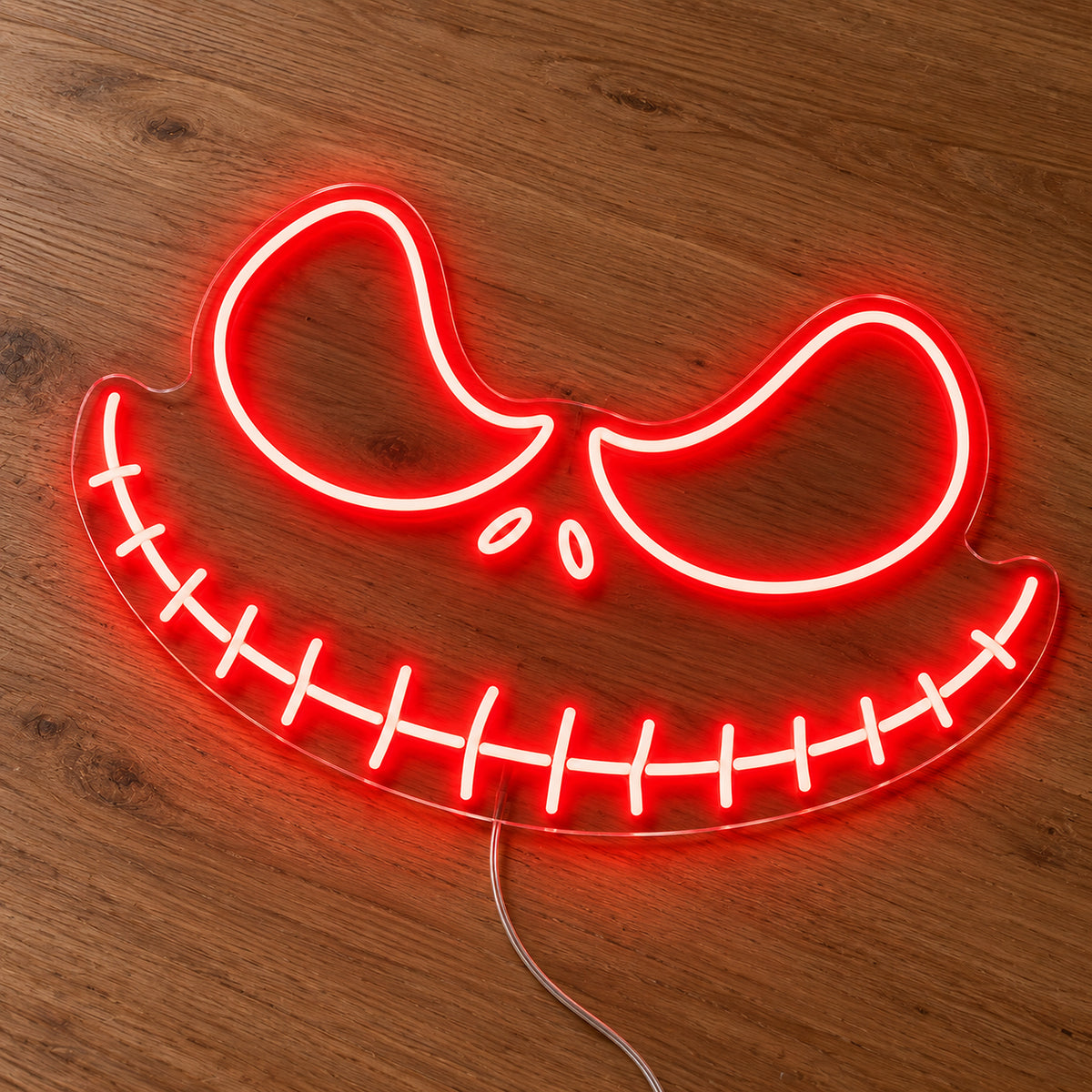 Red Jack Skellington Face Neon Sign featuring Jack Skellington's angled eyes and wide stitched grin in red, illuminated on clear acrylic lying on a natural oak wooden surface.