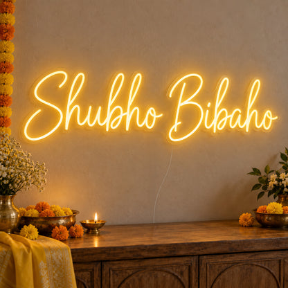 Shubho Bibaho Neon Sign on clear cut-to-shape acrylic, illuminated on a wall in a styled celebration setting