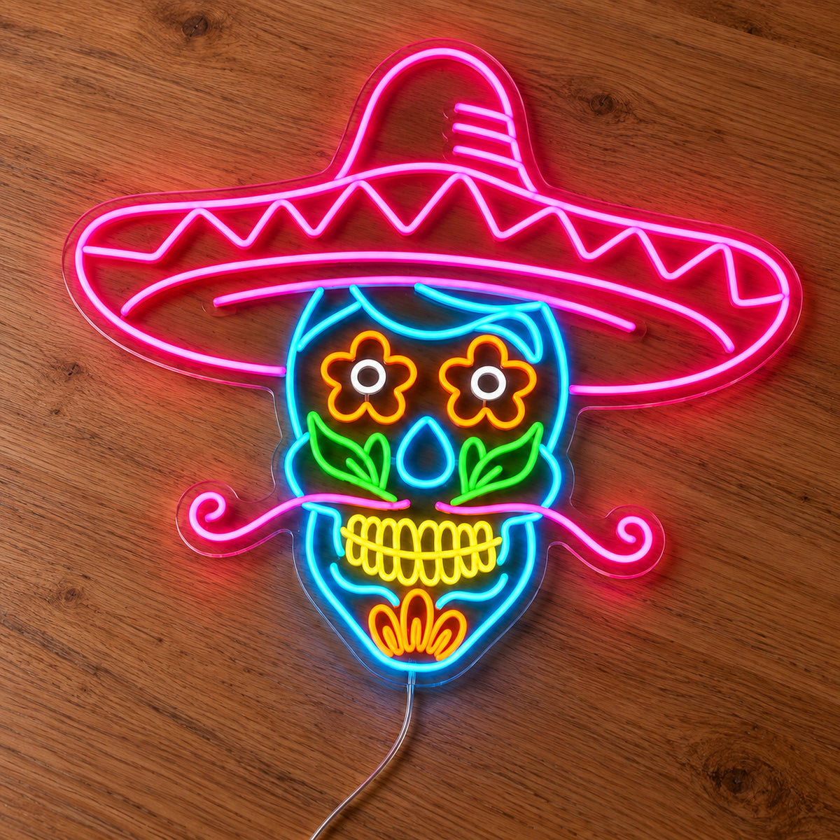 Sombrero Sugar Skull Neon Sign featuring a colourful sugar skull beneath a large pink sombrero, with floral eyes and a curled moustache, illuminated on clear acrylic lying on a natural oak wooden surface.