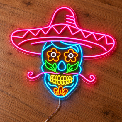 Sombrero Sugar Skull Neon Sign featuring a colourful sugar skull beneath a large pink sombrero, with floral eyes and a curled moustache, illuminated on clear acrylic lying on a natural oak wooden surface.