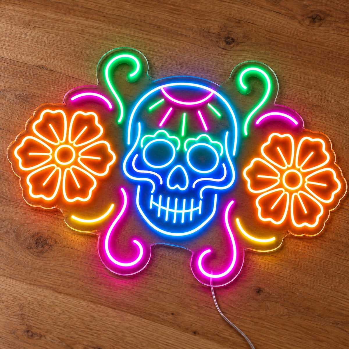 Sugar Skull with Flowers Neon Sign featuring a blue sugar skull flanked by orange flowers, with pink and green decorative curves, illuminated on clear acrylic lying on a natural oak wooden surface.