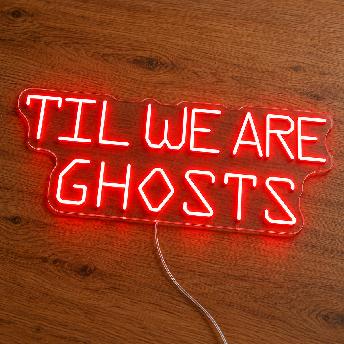 Til We Are Ghosts Block Letter Neon Sign featuring Til We Are Ghosts wording in red geometric block letters, illuminated on clear acrylic lying on a natural oak wooden surface.