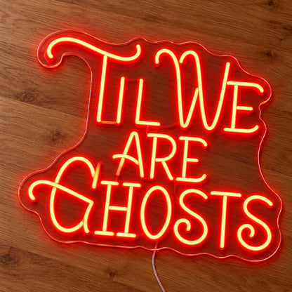 Til We Are Ghosts Ornate Neon Sign featuring Til We Are Ghosts wording in decorative orange lettering with curved flourishes, illuminated on clear acrylic lying on a natural oak wooden surface.