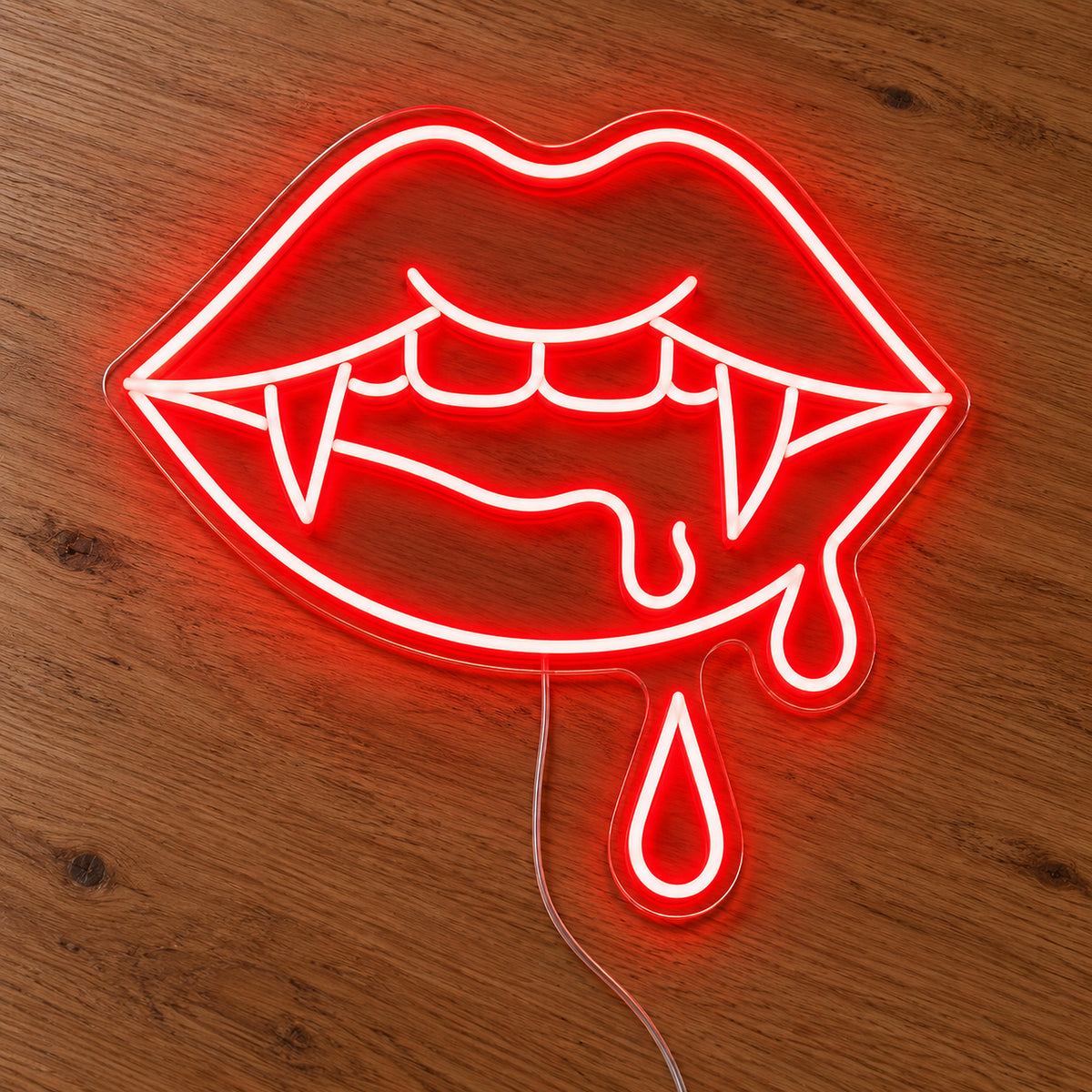 Vampire Lips with Double Blood Drops Neon Sign featuring red lips with pointed fangs and two hanging blood drops, illuminated on clear acrylic lying on a natural oak wooden surface.