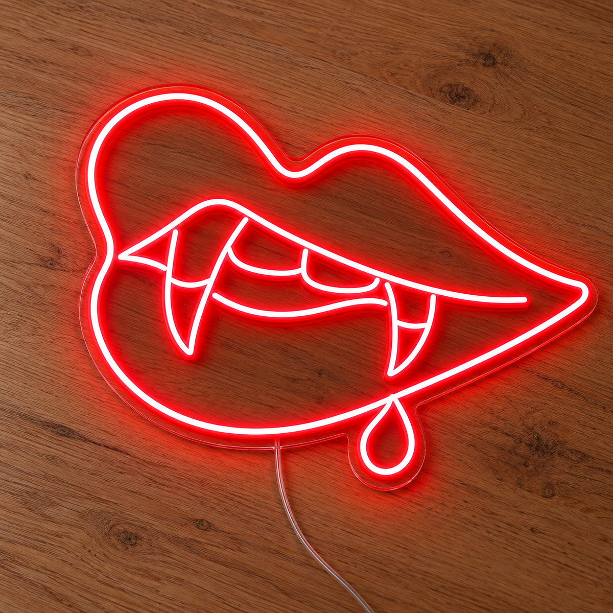 Vampire Lips with Single Blood Drop Neon Sign featuring red lips, pointed fangs and one hanging blood drop, illuminated on clear acrylic lying on a natural oak wooden surface.