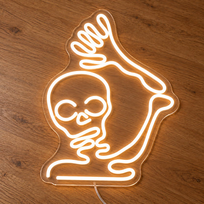 Waving Skeleton Neon Sign featuring a white skeleton with a raised arm and open hand, illuminated on clear acrylic lying on a natural oak wooden surface.
