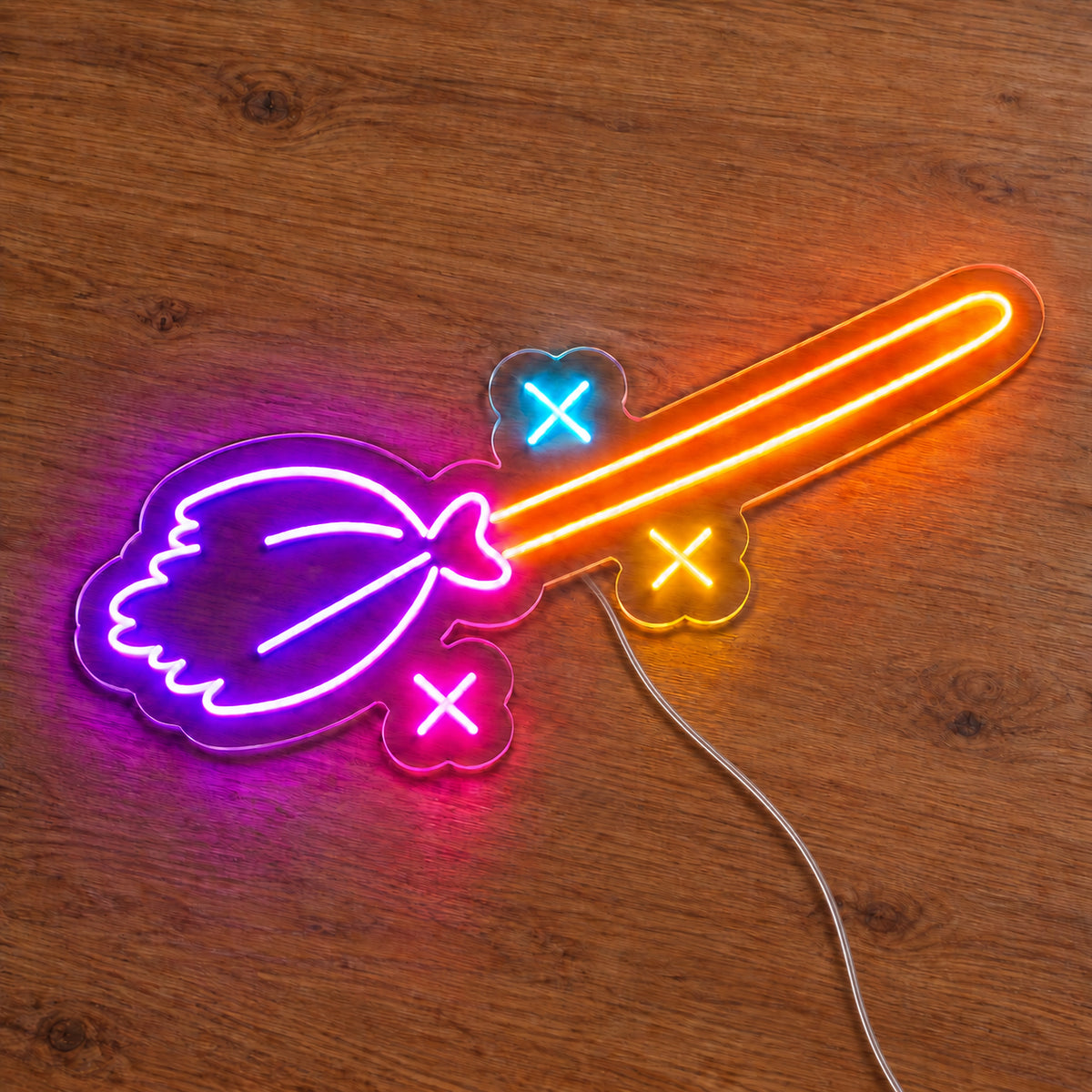 Witch Broom Neon Sign featuring a purple broom head, an orange handle and colourful sparkle details, illuminated on clear acrylic lying on a natural oak wooden surface.